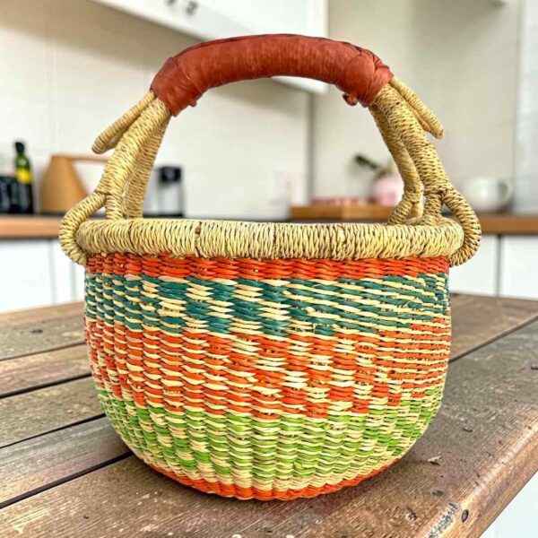 Small round Bolga basket with orange and green zigzag bands and a leather-wrapped handle, on a wooden table