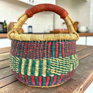 Small round Bolga basket with teal, red and natural upright stripes and a leather-wrapped handle, on a wooden table