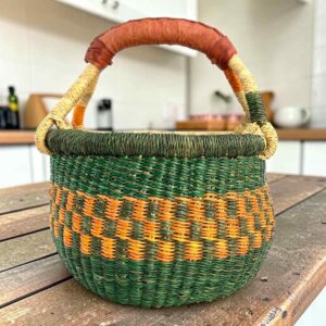 Small round Bolga basket with dark green bands around orange checks and a leather-wrapped handle, on a wooden table