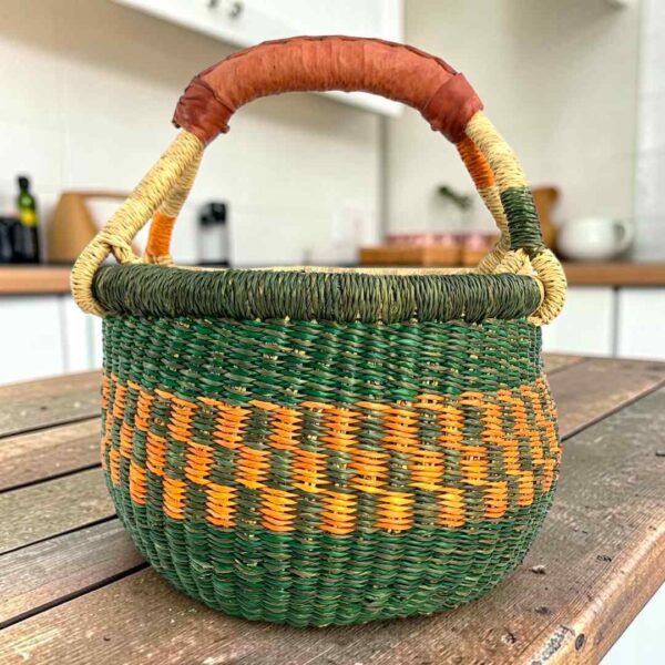 Small round Bolga basket with dark green bands around orange checks and a leather-wrapped handle, on a wooden table