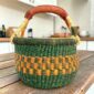 Small round Bolga basket with dark green bands around orange checks and a leather-wrapped handle, on a wooden table