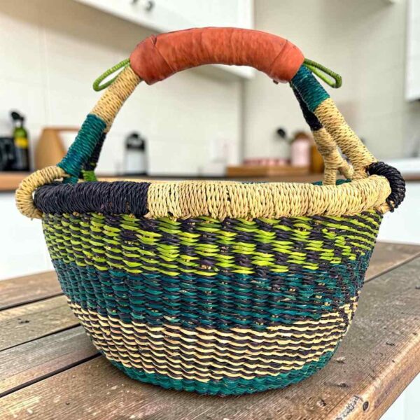 Small round Bolga basket with lime, teal and navy bands and a leather-wrapped handle, on a wooden table