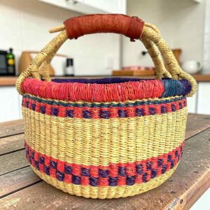 Small round Bolga basket with red and purple checked bands on natural straw and a leather-wrapped handle, on a wooden table
