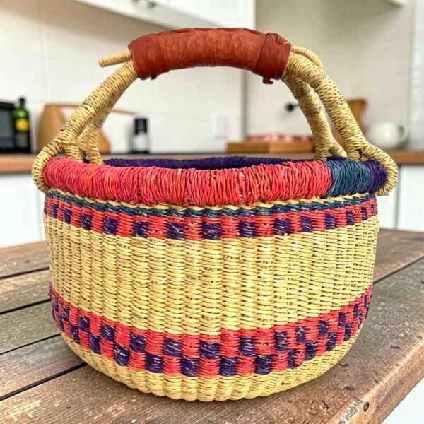 small-round-basket-731-downstream-red-purple-bolga-african-basket-bashiri-main Small round Bolga basket with red and purple checked bands on natural straw and a leather-wrapped handle, on a wooden table