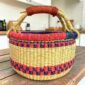 Small round Bolga basket with red and purple checked bands on natural straw and a leather-wrapped handle, on a wooden table
