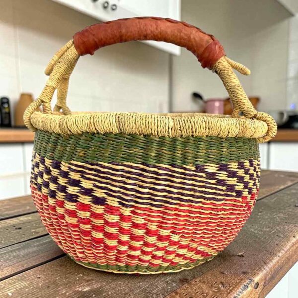 Small round Bolga basket with red checks under green and navy bands and a leather-wrapped handle, on a wooden table