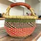 Small round Bolga basket with red checks under green and navy bands and a leather-wrapped handle, on a wooden table