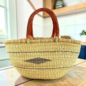 Natural Mega Wawa Bolga basket with fine black lines, a small diamond motif and two leather handles, on a wooden table