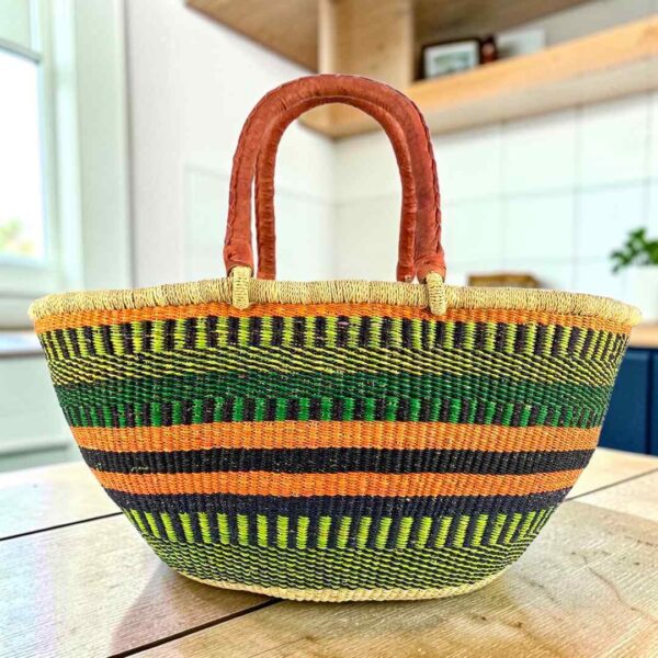 Green and orange striped Mega Wawa Bolga basket with lime and navy bands and two leather handles, on a wooden table