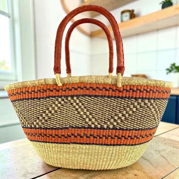 wawa-basket-262-occo-main Mega Wawa Bolga basket with orange bands, a black and natural zigzag panel and two leather handles, on a wooden table