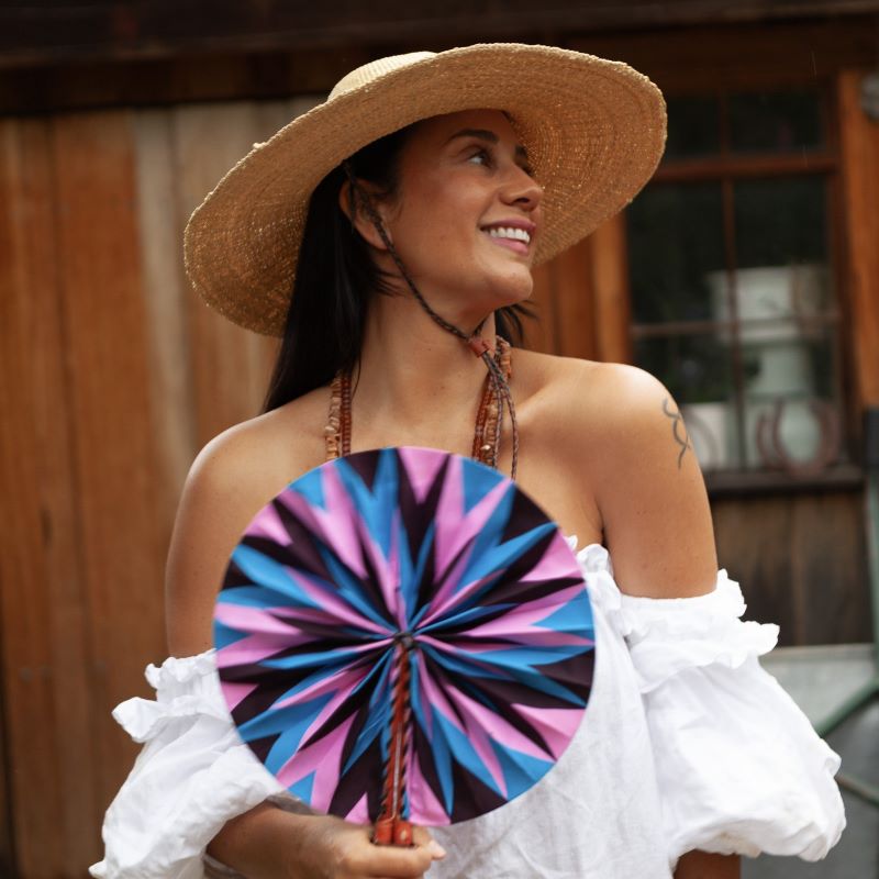 A woman wearing woven straw hat in natural colour, holding a fabric fan and smiling.